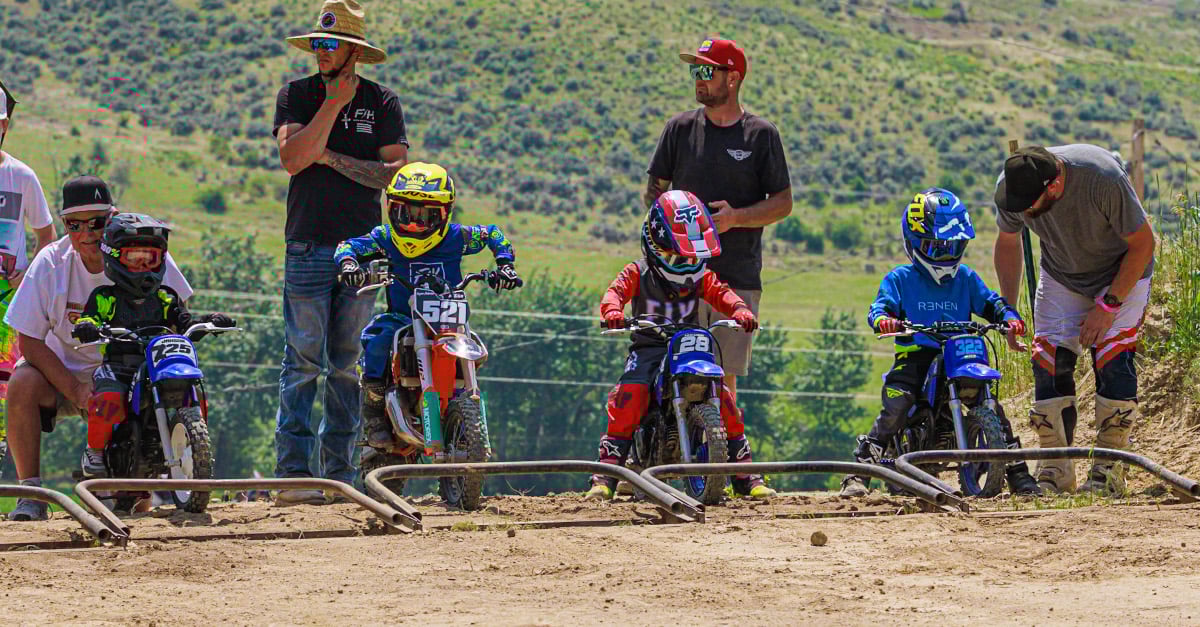 Children lined up ready to race motorbikes with adults standing next to them by the gate